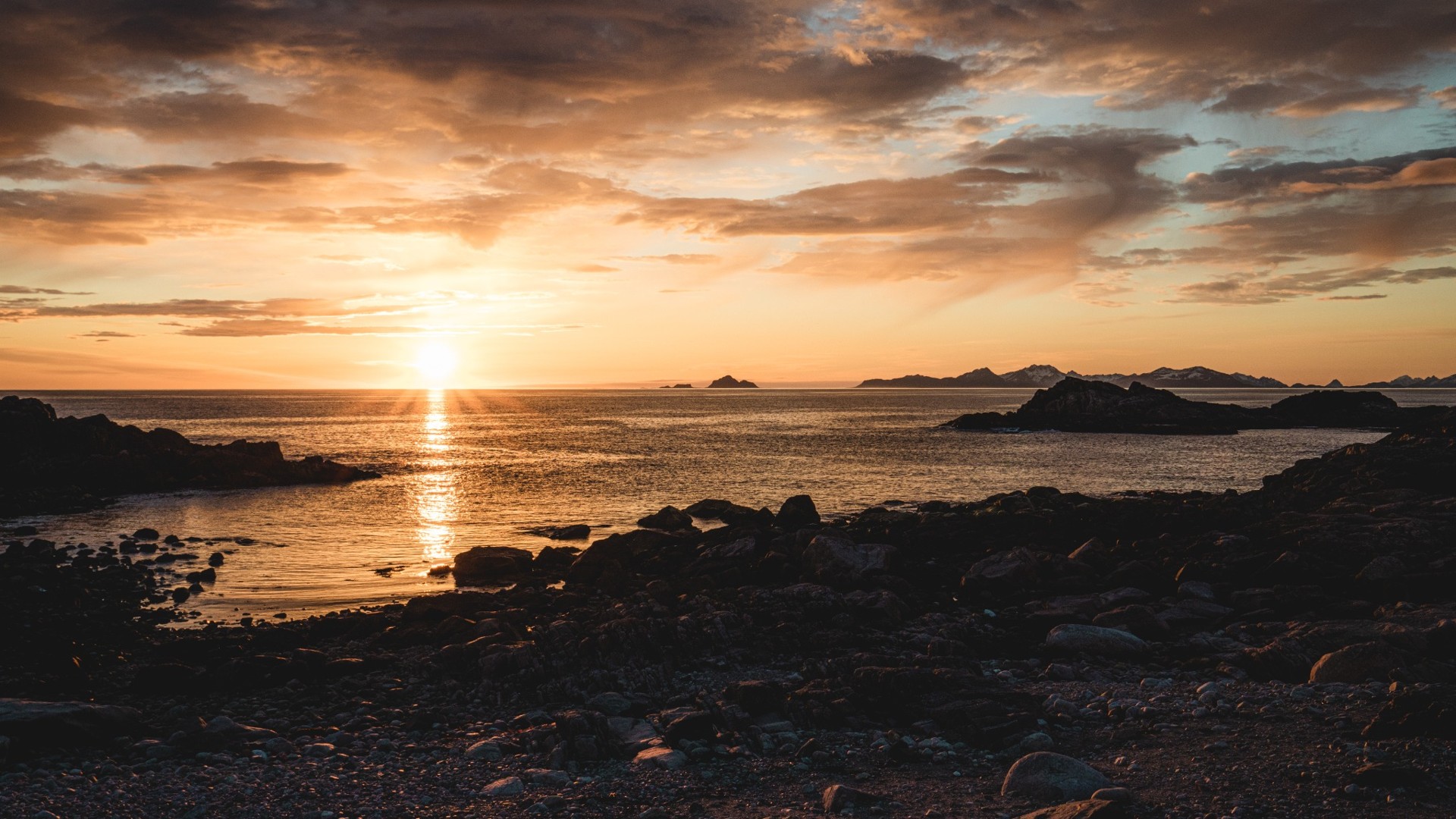 Wide cinematic sunset over the Arctic sea with sunbeams, rocky shore and distant Lofoten peaks