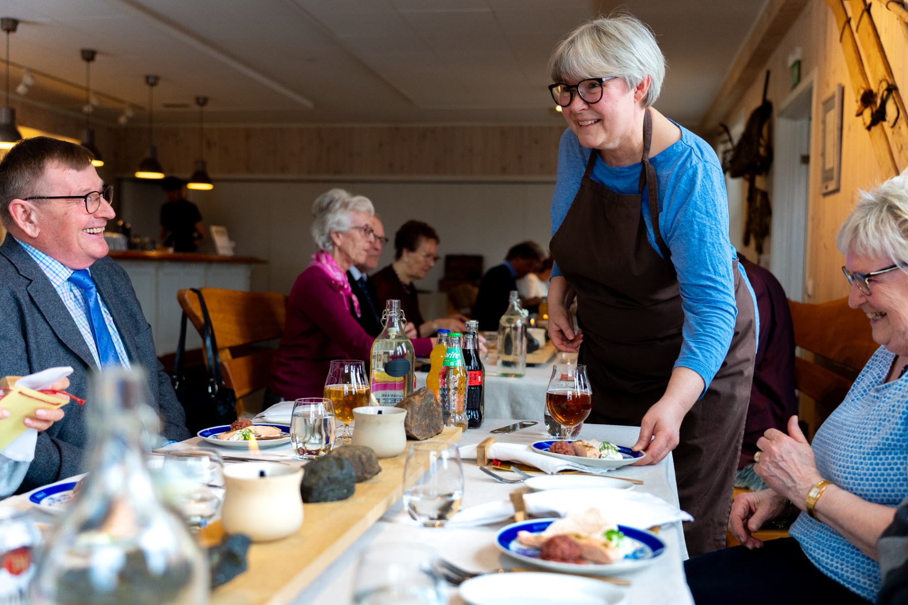 Warm host in apron laughing with seated guests as she serves plated courses