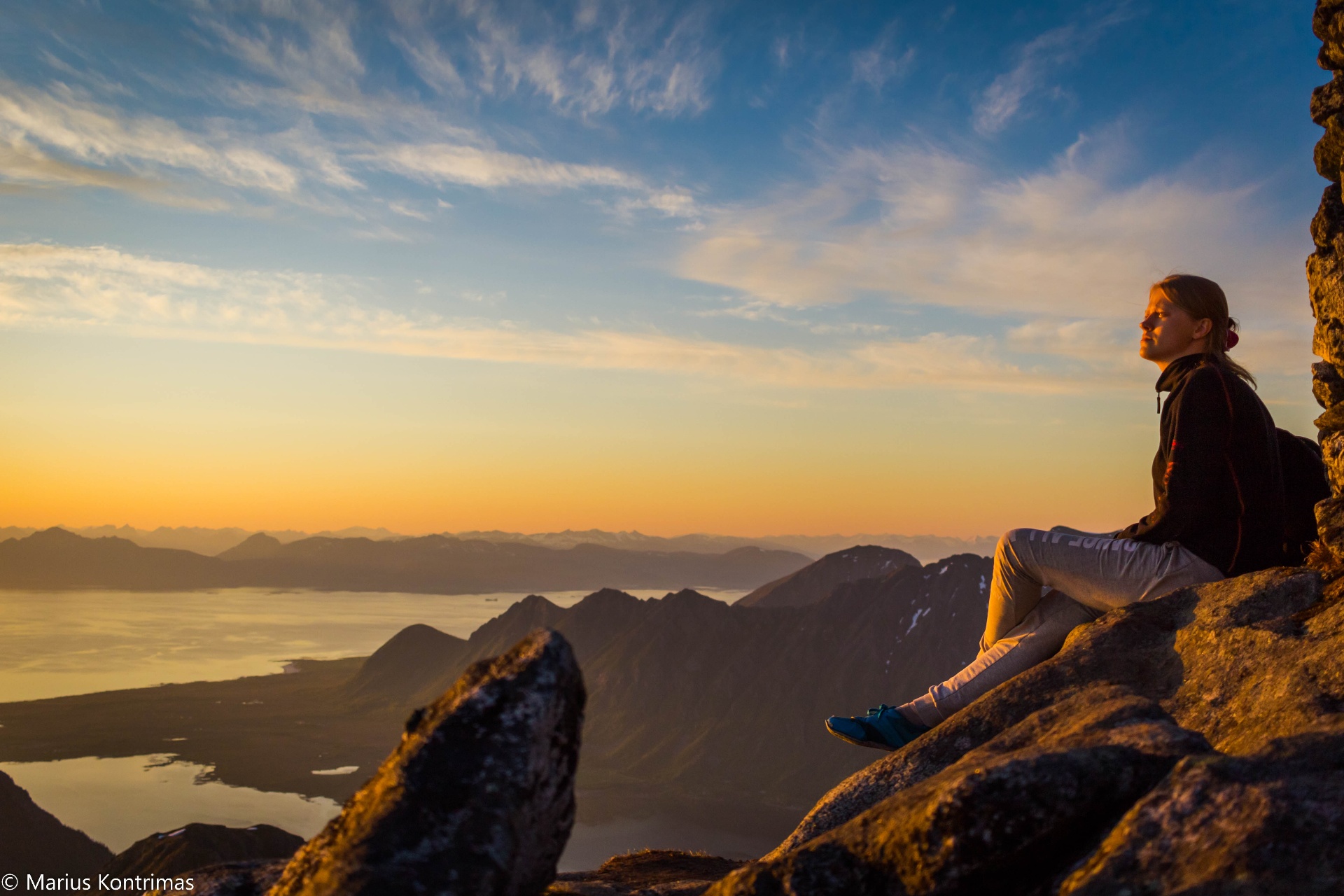 Guest seated on a mountain ledge in warm midnight-sun light above a sweeping Lofoten fjord panorama