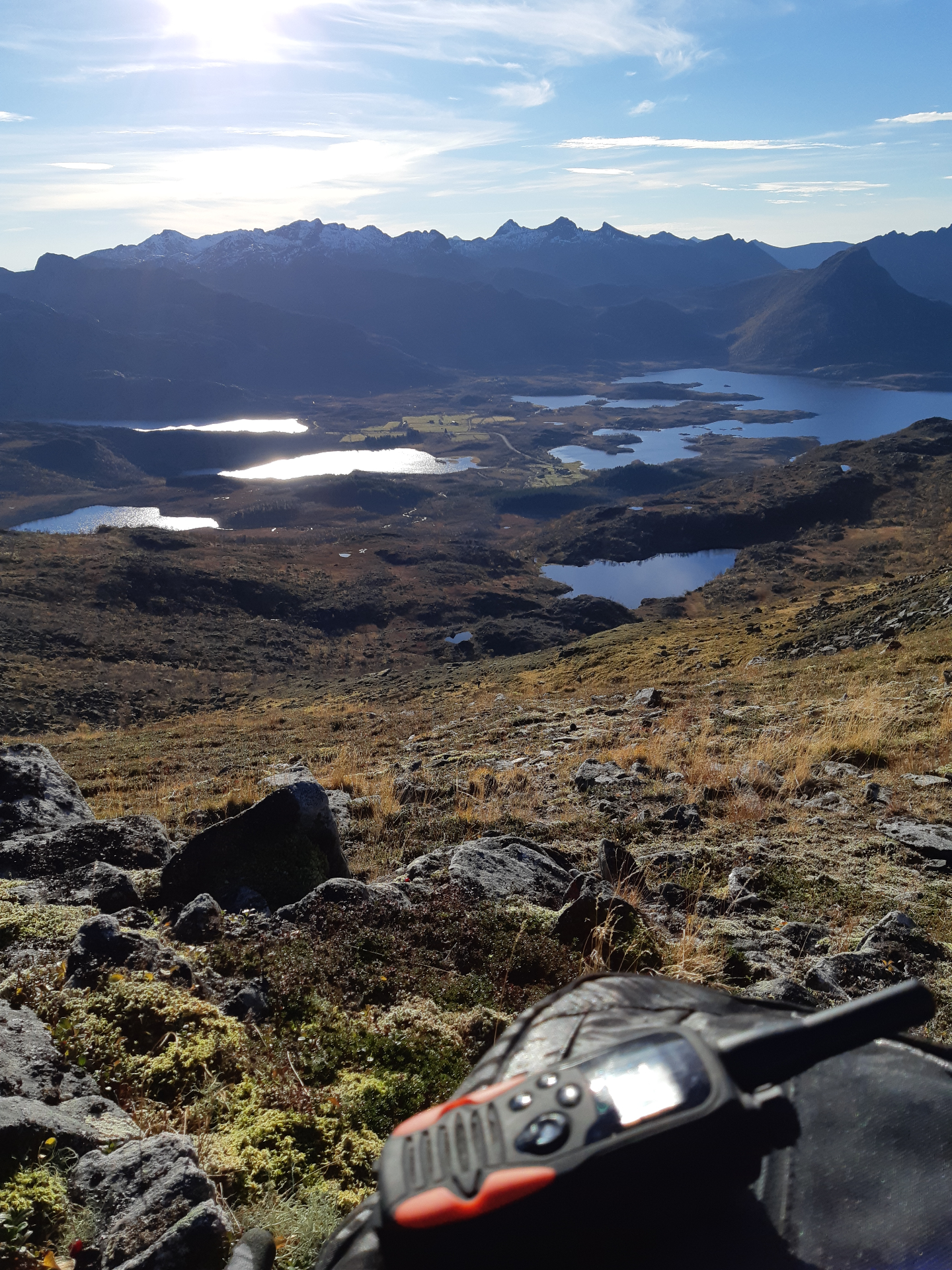 Handheld radio on mossy rocks at a summit overlooking Lofoten lakes and jagged peaks