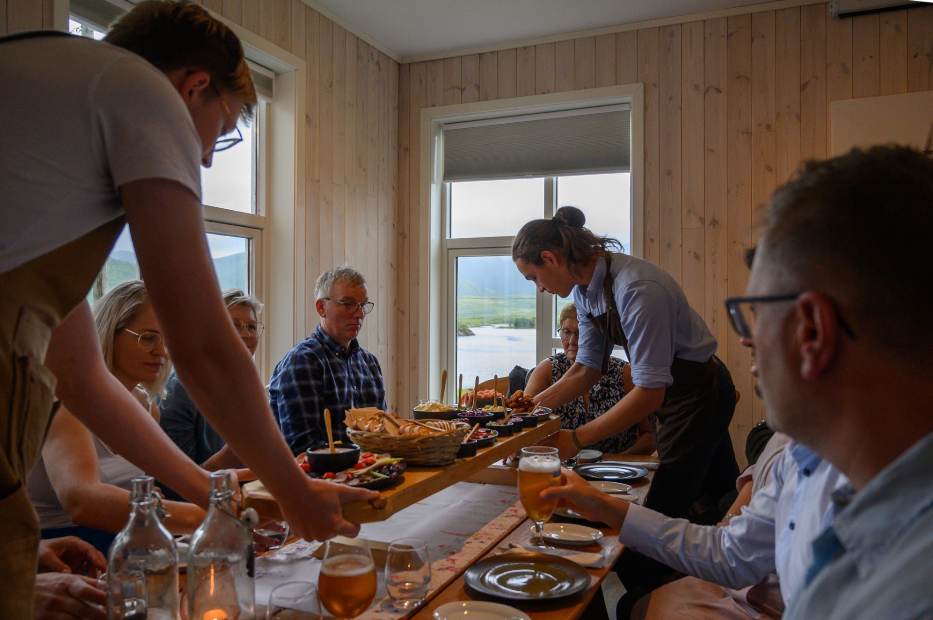 Hosts serving a shared tasting spread to guests around a long wooden table