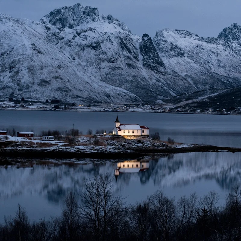 Sildpollnes church lit at blue hour on a snowy headland, mirrored in the still fjord below snow-covered peaks