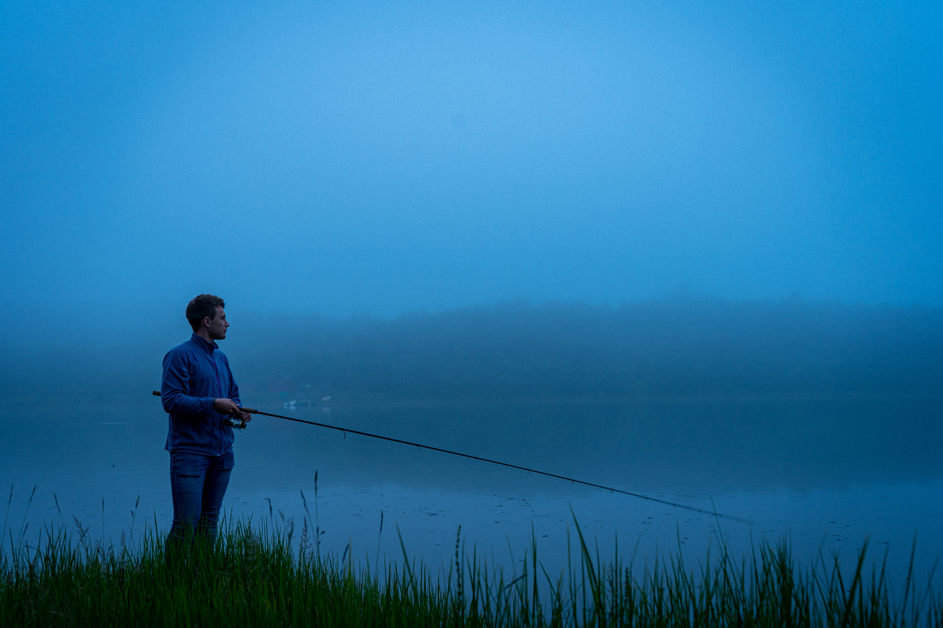 Lone angler casting into misty Arctic water during deep blue summer twilight