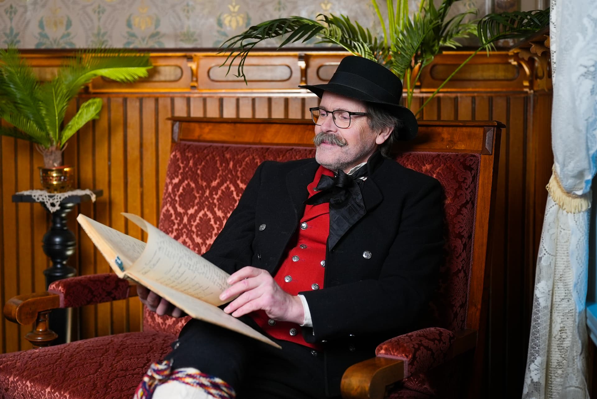 Storyteller in black bunad and red vest reading an old handwritten ledger in a heritage parlour