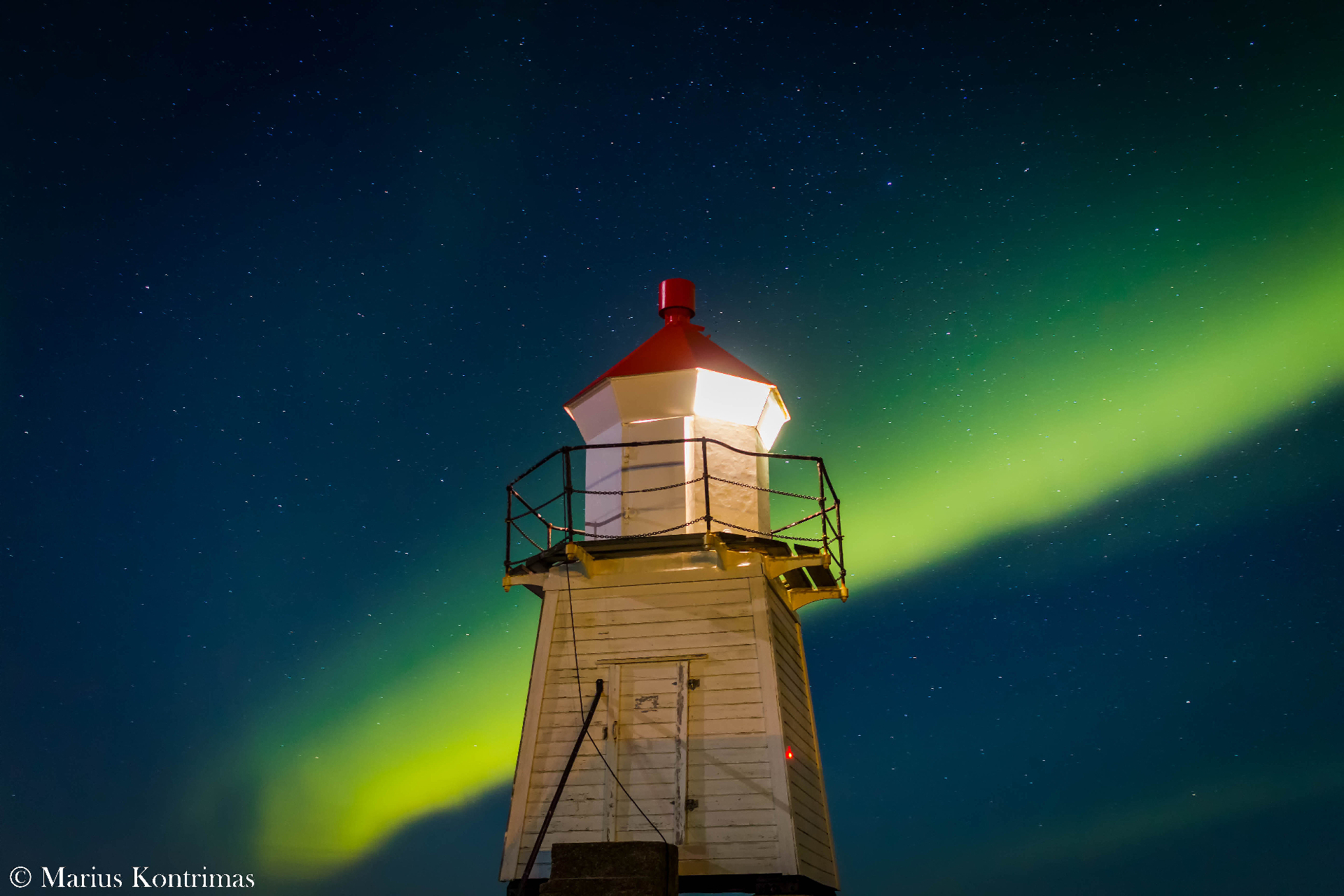 Illuminated lighthouse beneath a green aurora over a starlit Arctic sky