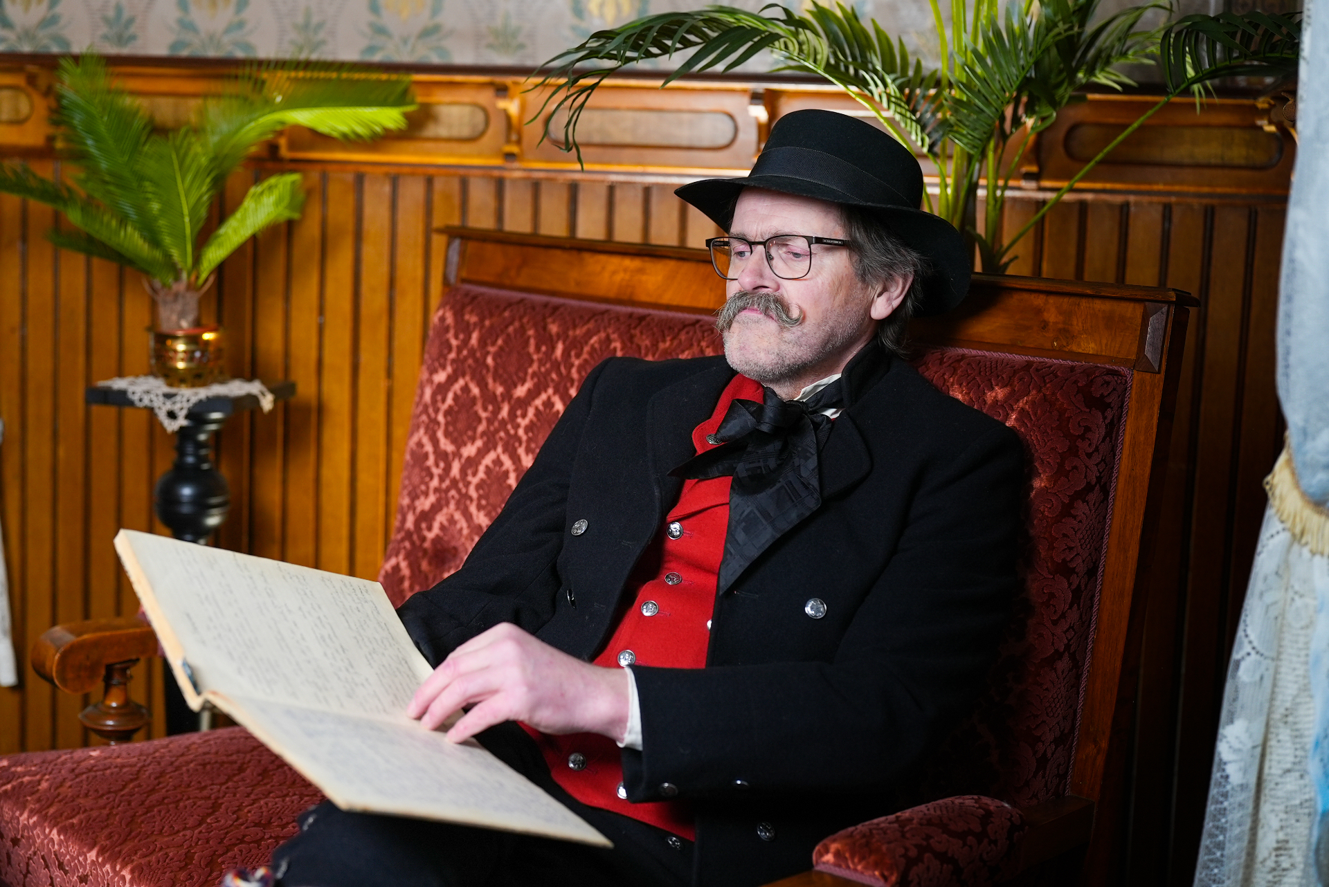 Storyteller in black hat and red bunad vest reading from an old handwritten ledger