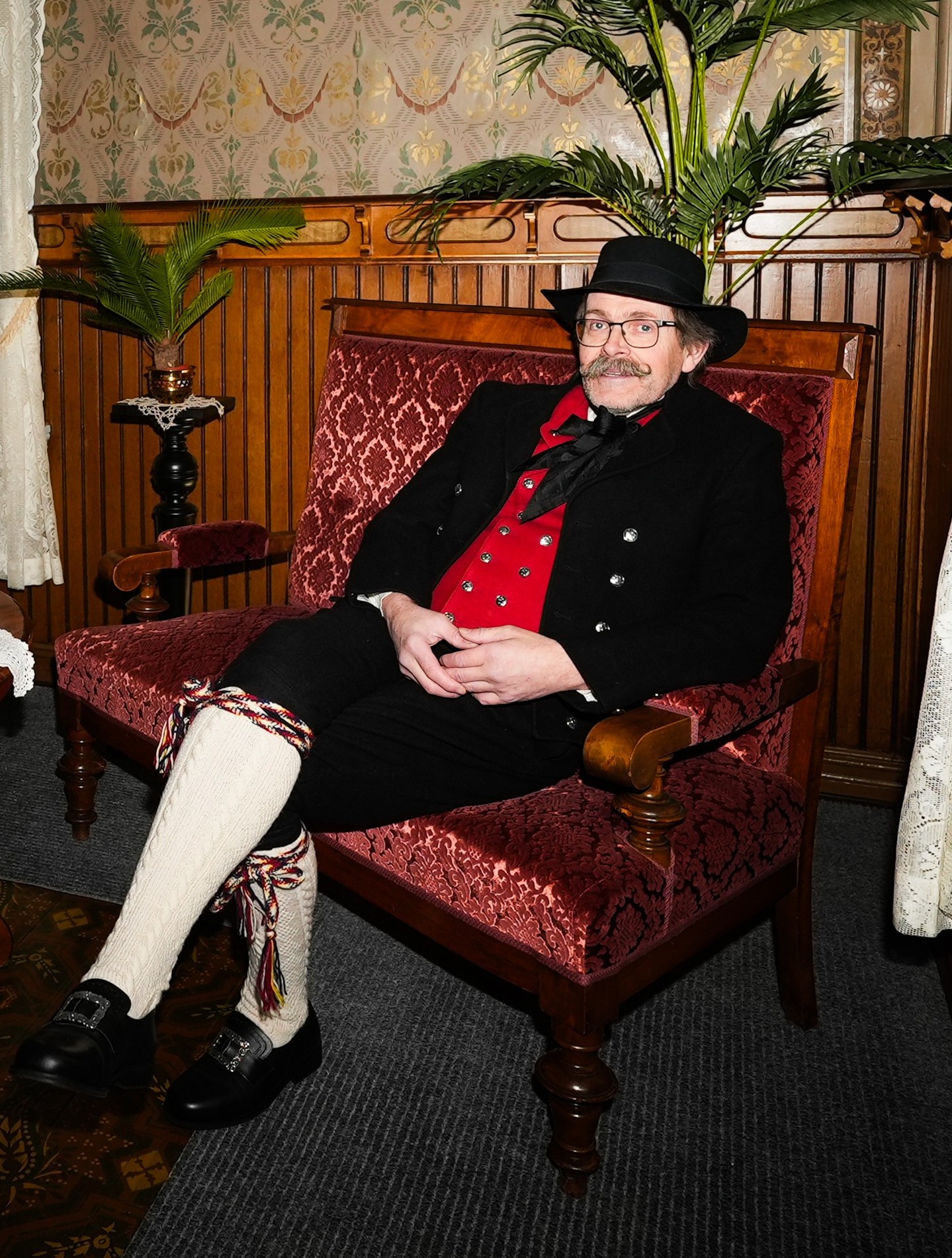 Storyteller Roy in traditional Nordland bunad seated in a crimson velvet chair inside a historic parlour