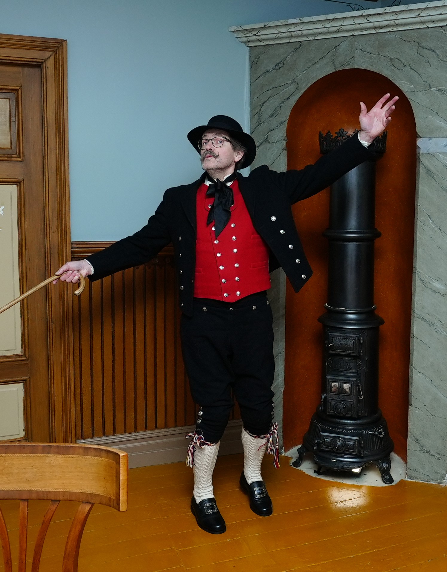 Storyteller Roy in traditional Nordland bunad gesturing beside a black cast-iron stove in a heritage parlour