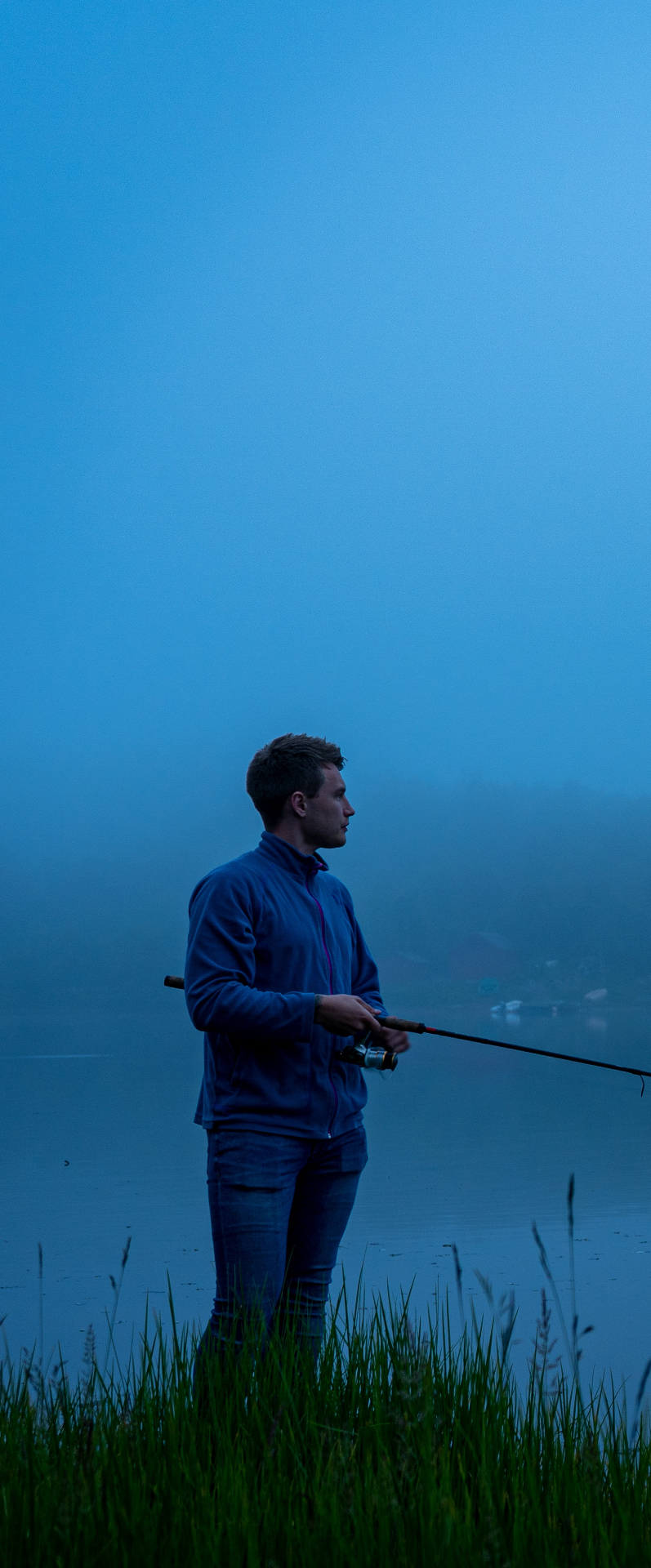 Young angler standing in tall grass by a misty blue-hour lake in Lofoten