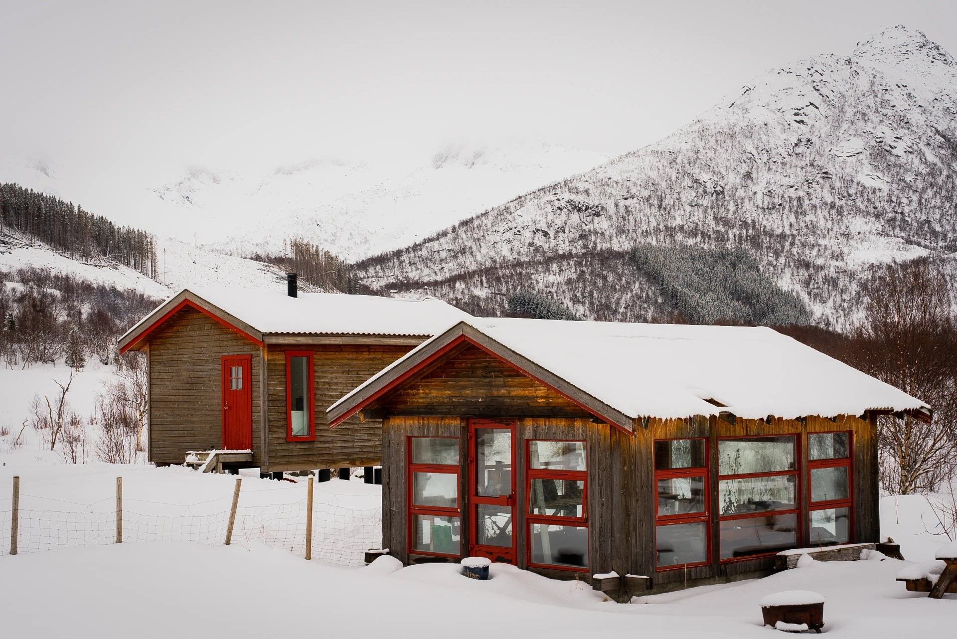 Two red-framed timber cabins under deep snow with Lofoten peaks rising behind