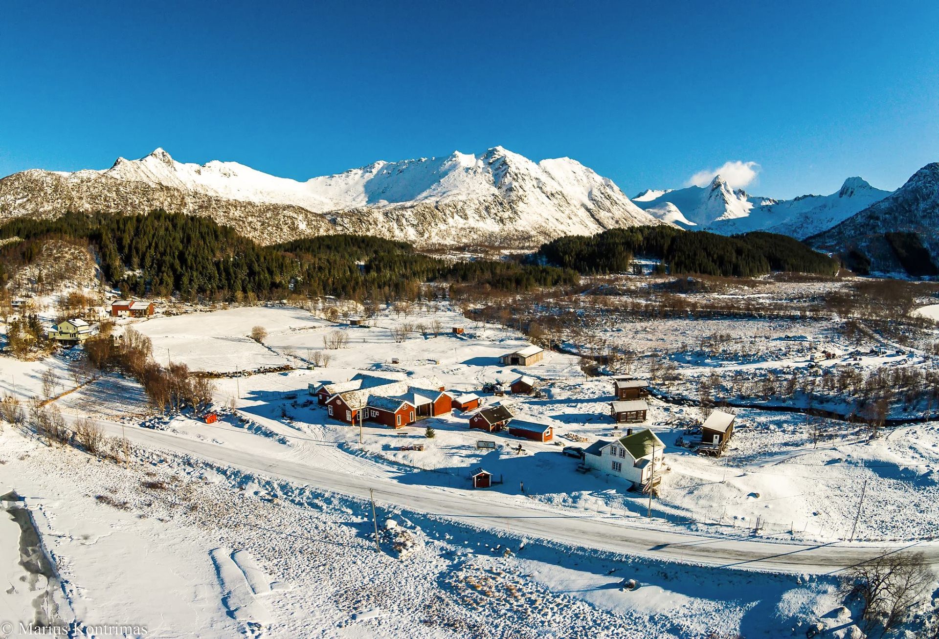 Aerial view of a red-cabin Lofoten farmstead in fresh snow beneath sharp winter peaks