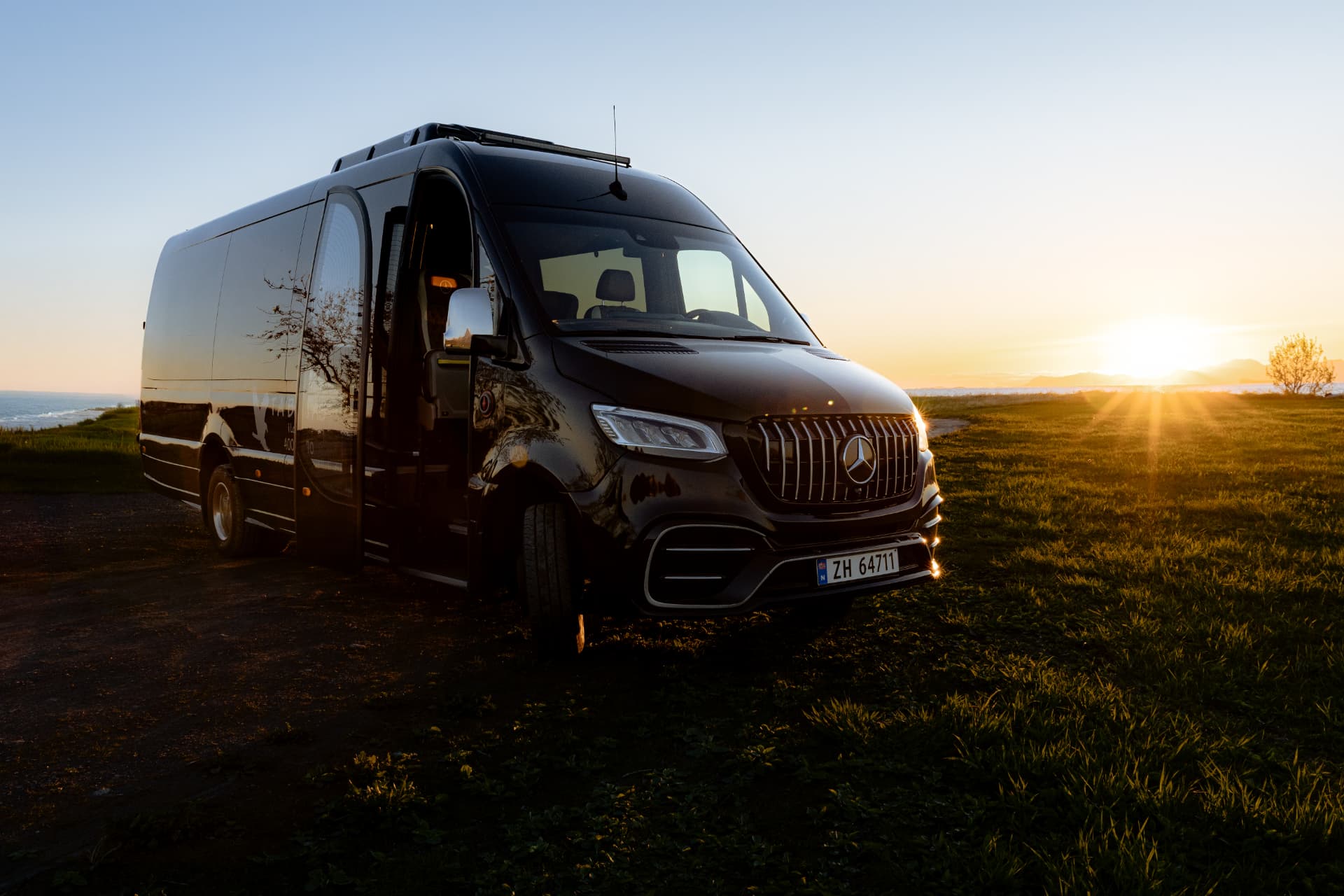 Black Mercedes-Benz minibus parked on a coastal meadow at Livland Gård with the low Arctic sun flaring across the sea