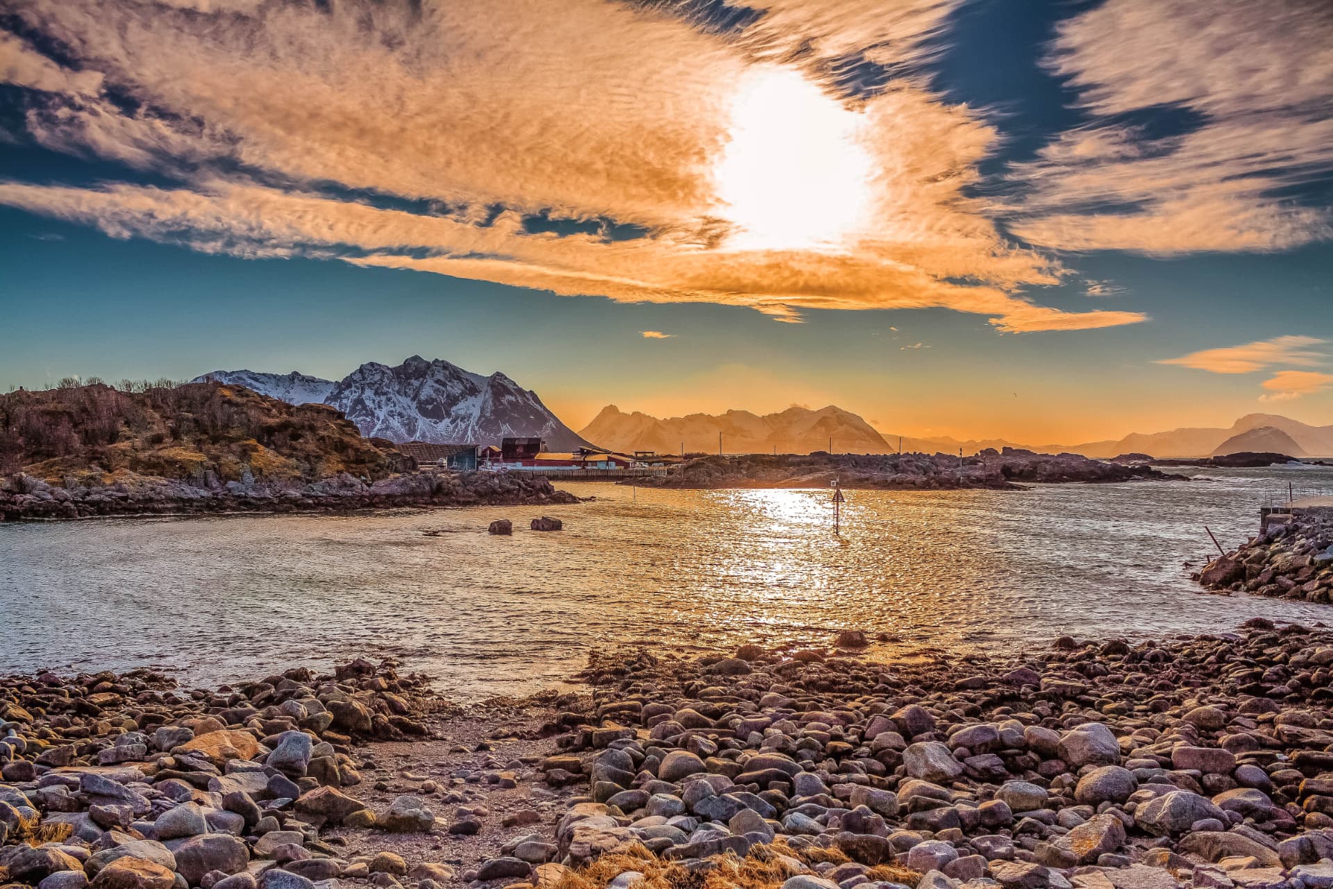 Low Arctic sun burning through cloud above the rocky shoreline at Laukvik with snow-capped peaks beyond
