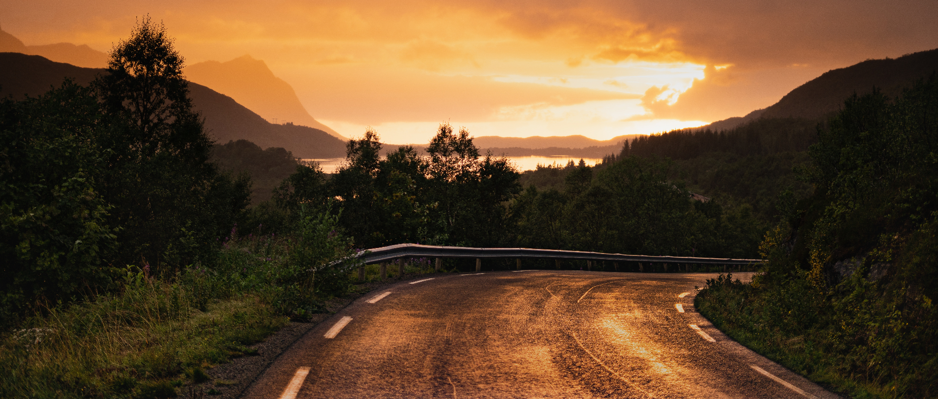 Empty Lofoten road curving toward a molten copper sunset over fjord and mountains
