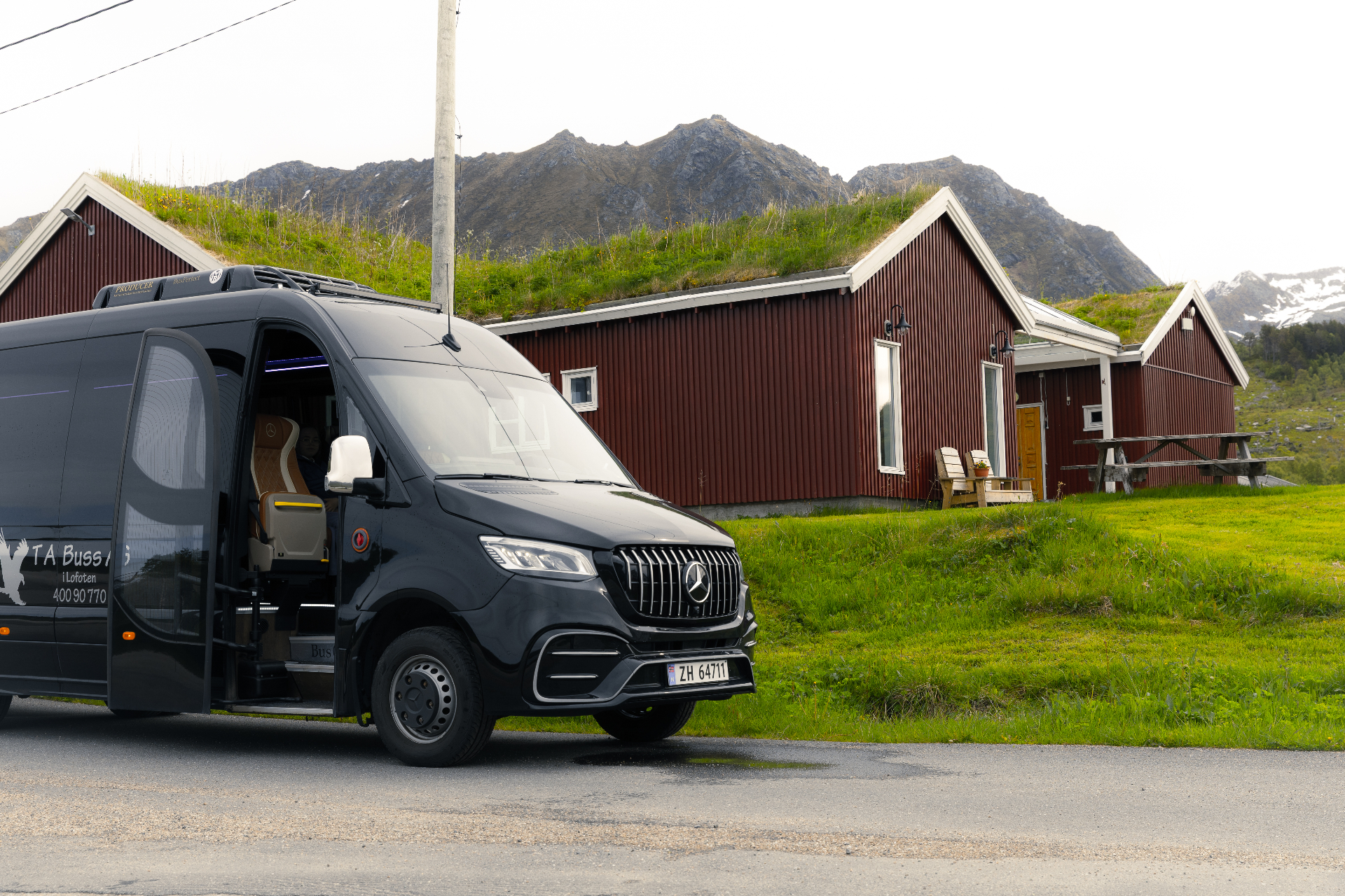 Black Mercedes Sprinter parked beside red turf-roof cabins under a Lofoten peak