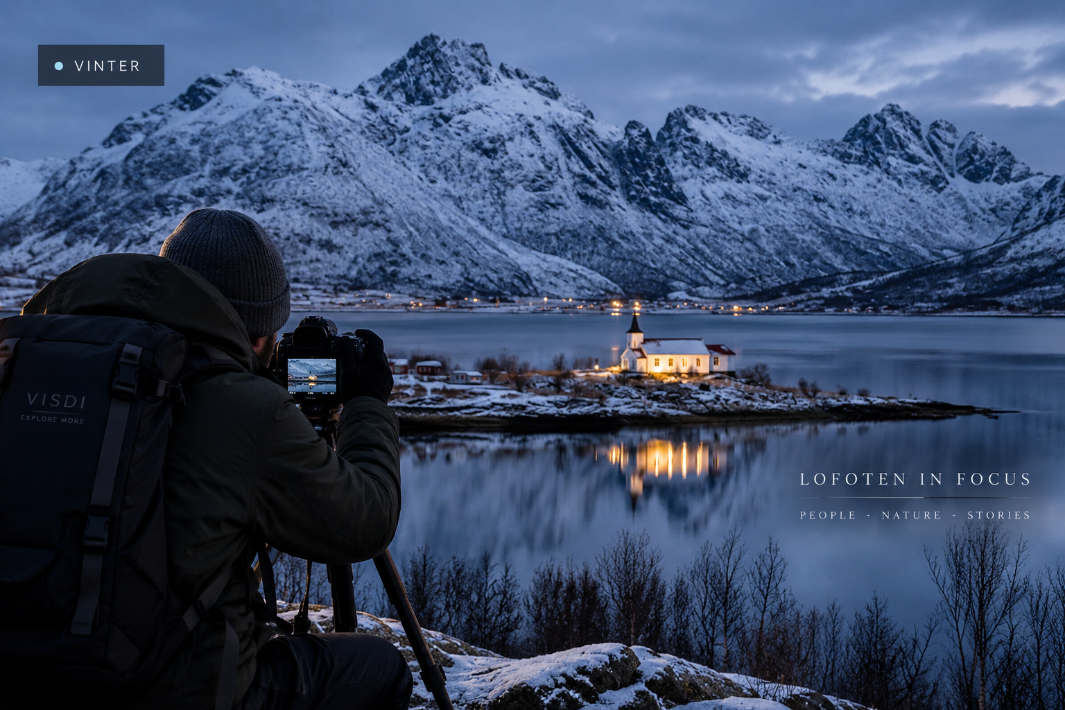 A photographer looks across the water towards a lit church and snow-covered Lofoten peaks at dusk