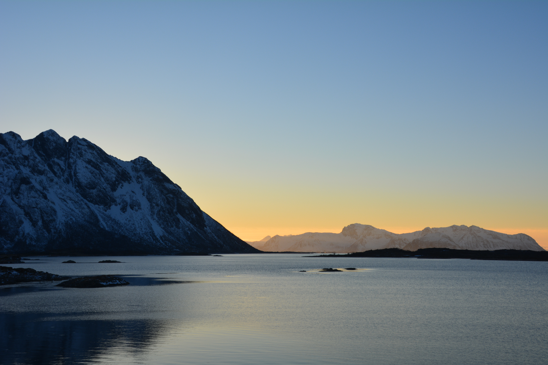 Snow-clad Lofoten mountain silhouetted against a soft copper dawn over a still fjord
