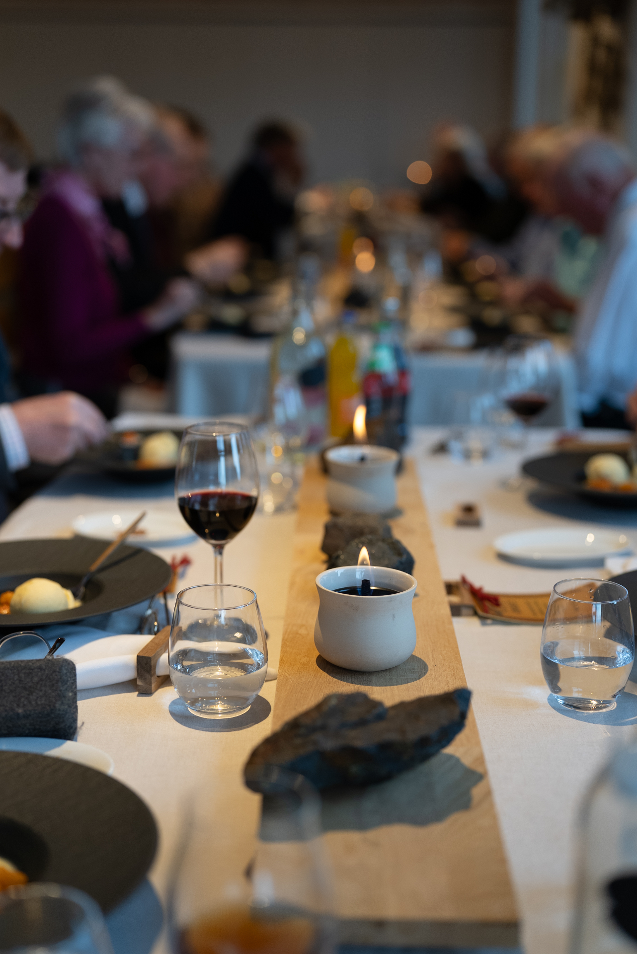 Long table set with wine, water glasses, candlelight and slate details — guests dining softly in the background