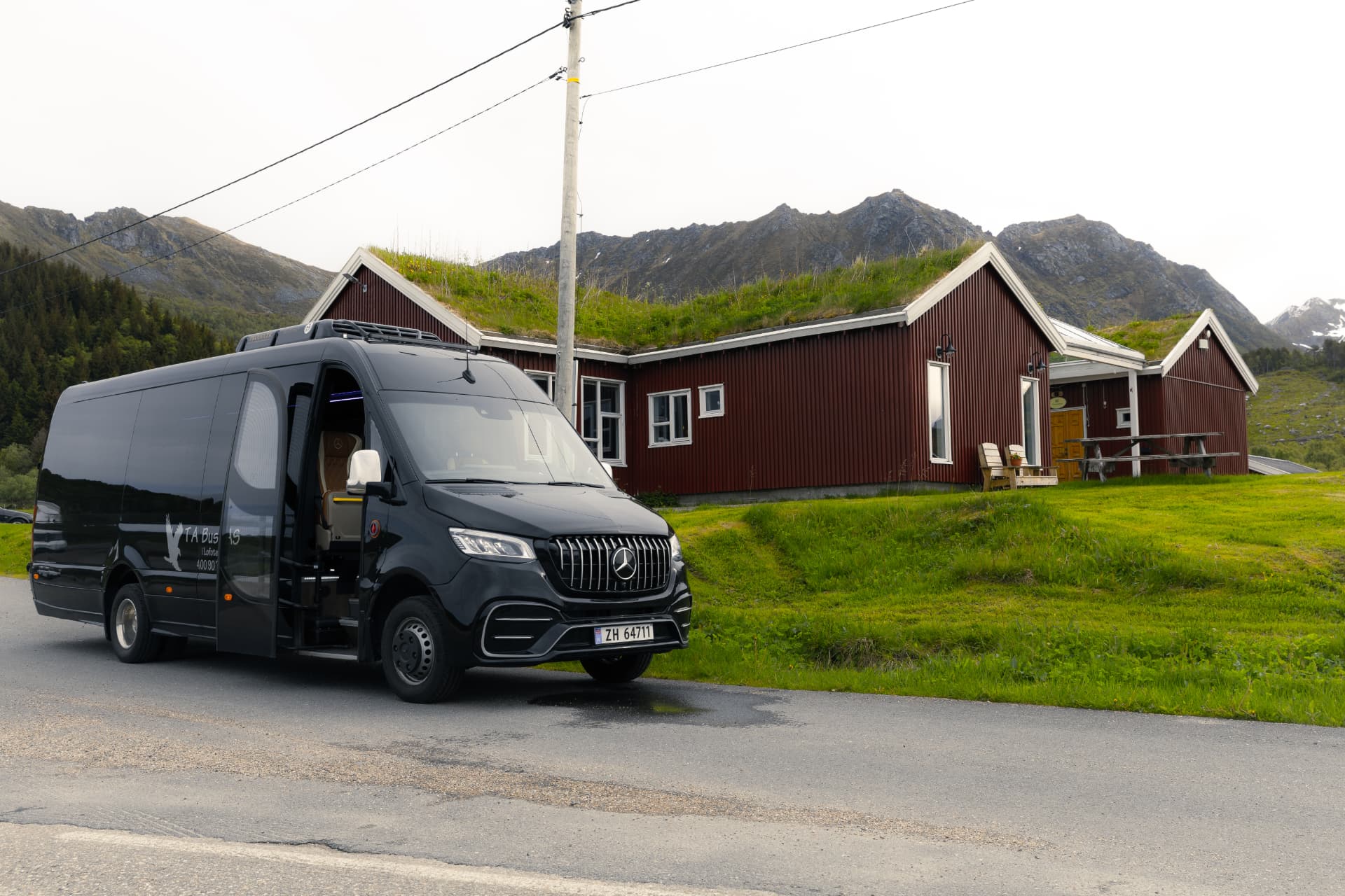 Black TA Buss Mercedes-Benz Sprinter parked by the red turf-roof cabins at Livland Gård with Lofoten peaks behind