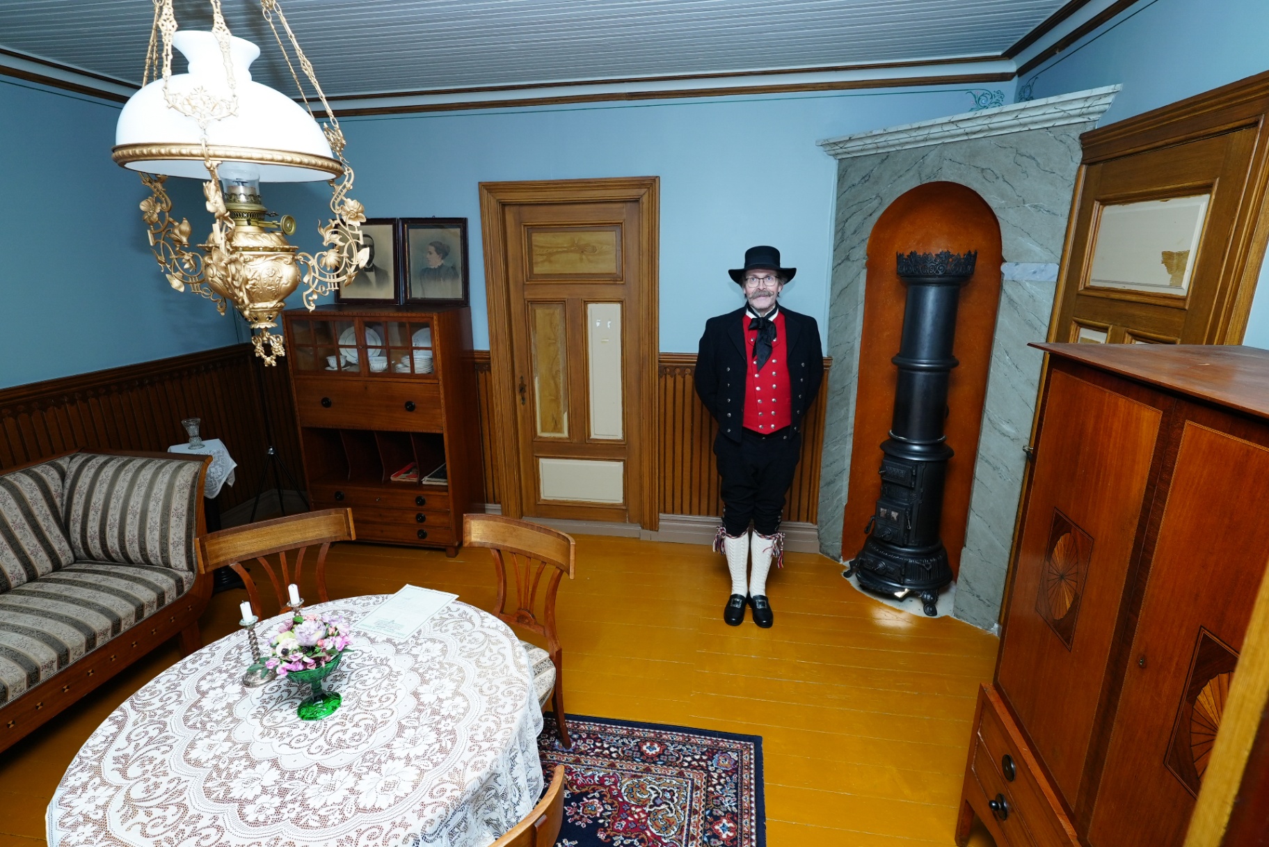 Bunad-clad guide standing in a restored blue heritage parlour with brass lamp and wood stove